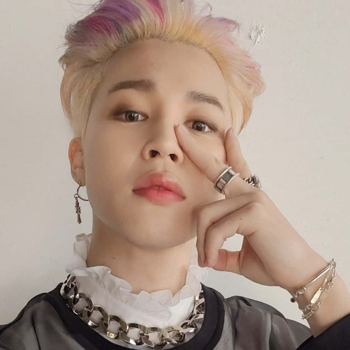 BTS 成員 Jimin 在拍攝中展現魅力 壁紙 — 一個擁有粉紅色和黃色頭髮的人，穿著白色荷葉邊領和粗銀鏈項鍊，將下巴擱在手上。