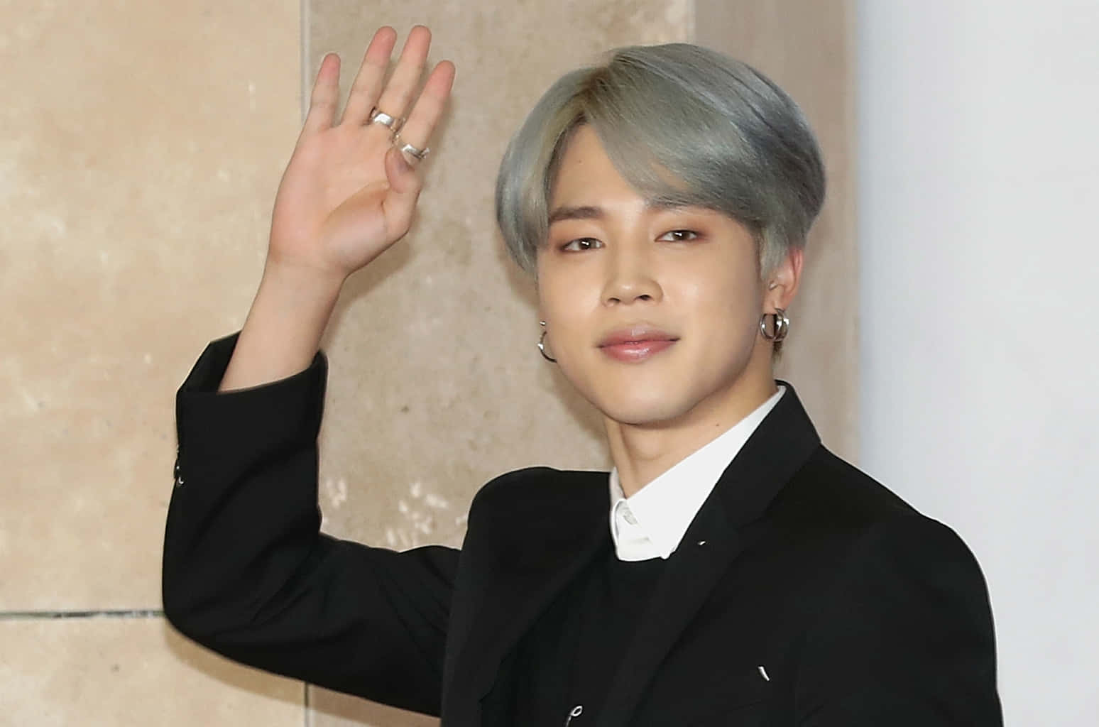 BTS 成員 Jimin 笑容自信滿滿 壁紙 — 一位銀髮年輕男子穿著黑色西裝和白色襯衫，在米色瓷磚牆前愉快地揮手。