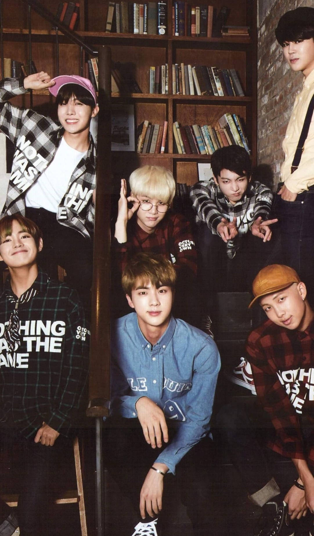 BTS 手機顯示畫面：圖書館閱讀風景 壁紙 — 七個年輕男子穿著休閒格仔和圖案T恤，在一個有木書架和磚牆的舒適圖書館裡頑皮地擺姿勢。