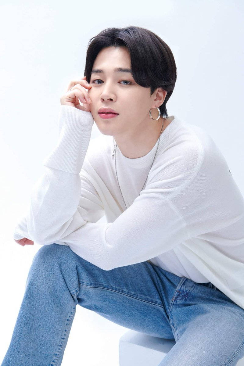 BTS 朴智旻 Chilsung代言 壁紙 — 一位深色頭髮、戴著耳環的年輕男子，穿著白色毛衣和牛仔褲，隨意地坐在明亮的白色背景前。