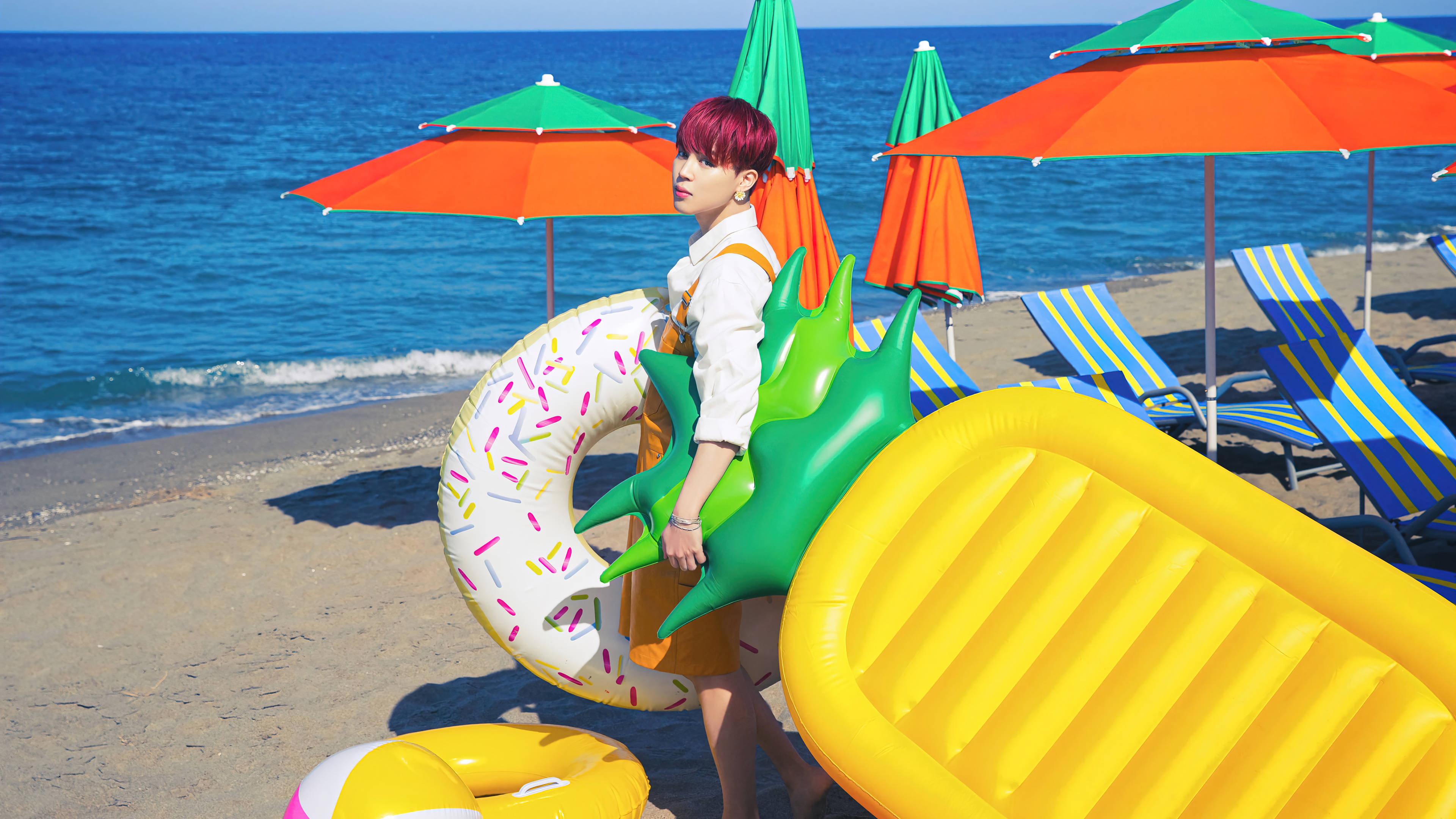 BTS 朴智旻 海灘 壁紙 — 一個粉紅色頭髮的人站在陽光明媚的海灘上，在彩色遮陽傘和躺椅附近拿著巨大的充氣玩具。