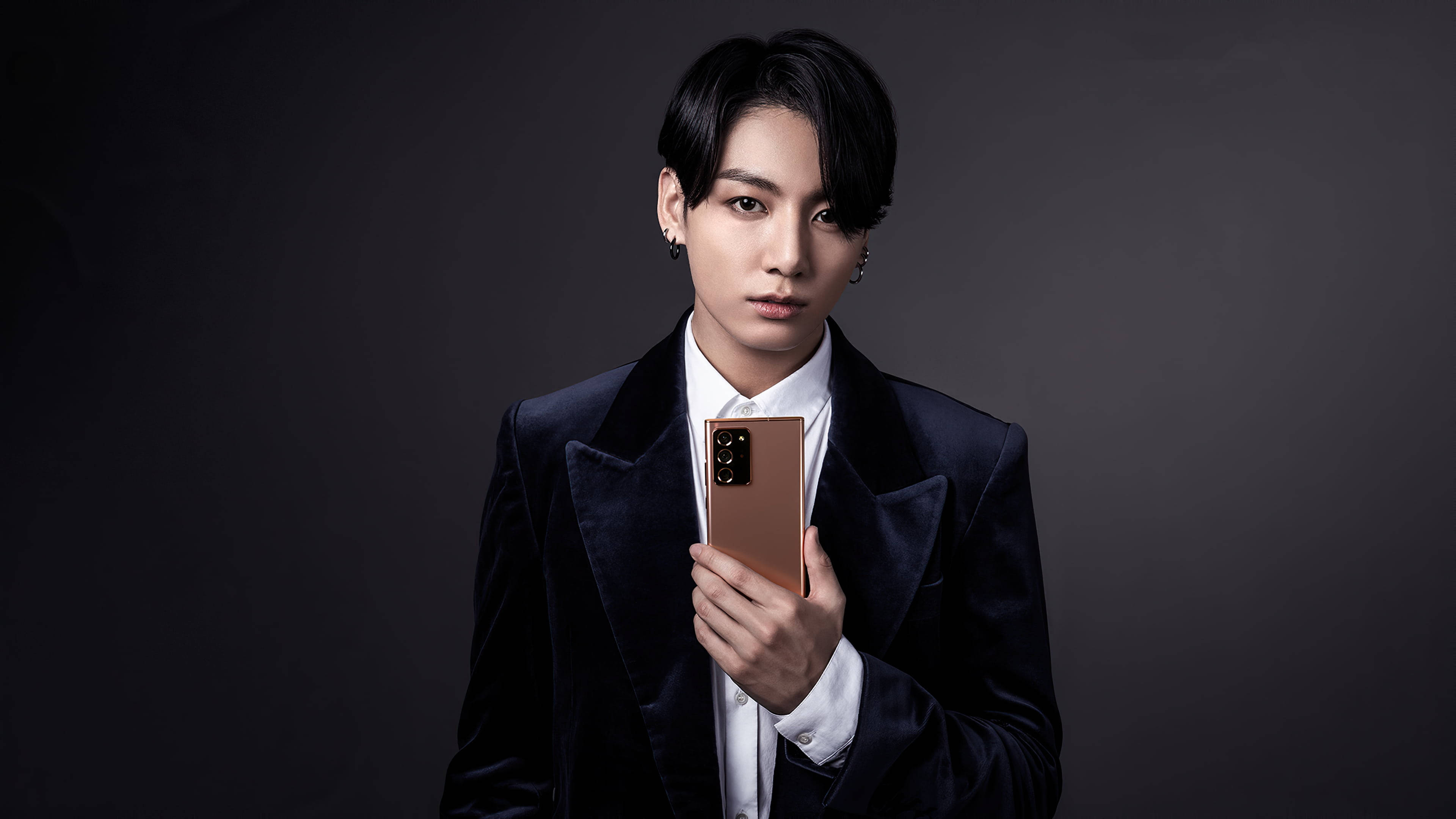 BTS 田柾國 可愛 Samsung 壁紙 — 一位身穿深色天鵝絨西裝的年輕男子，在陰鬱的灰色背景前拿著玫瑰金智能手機。