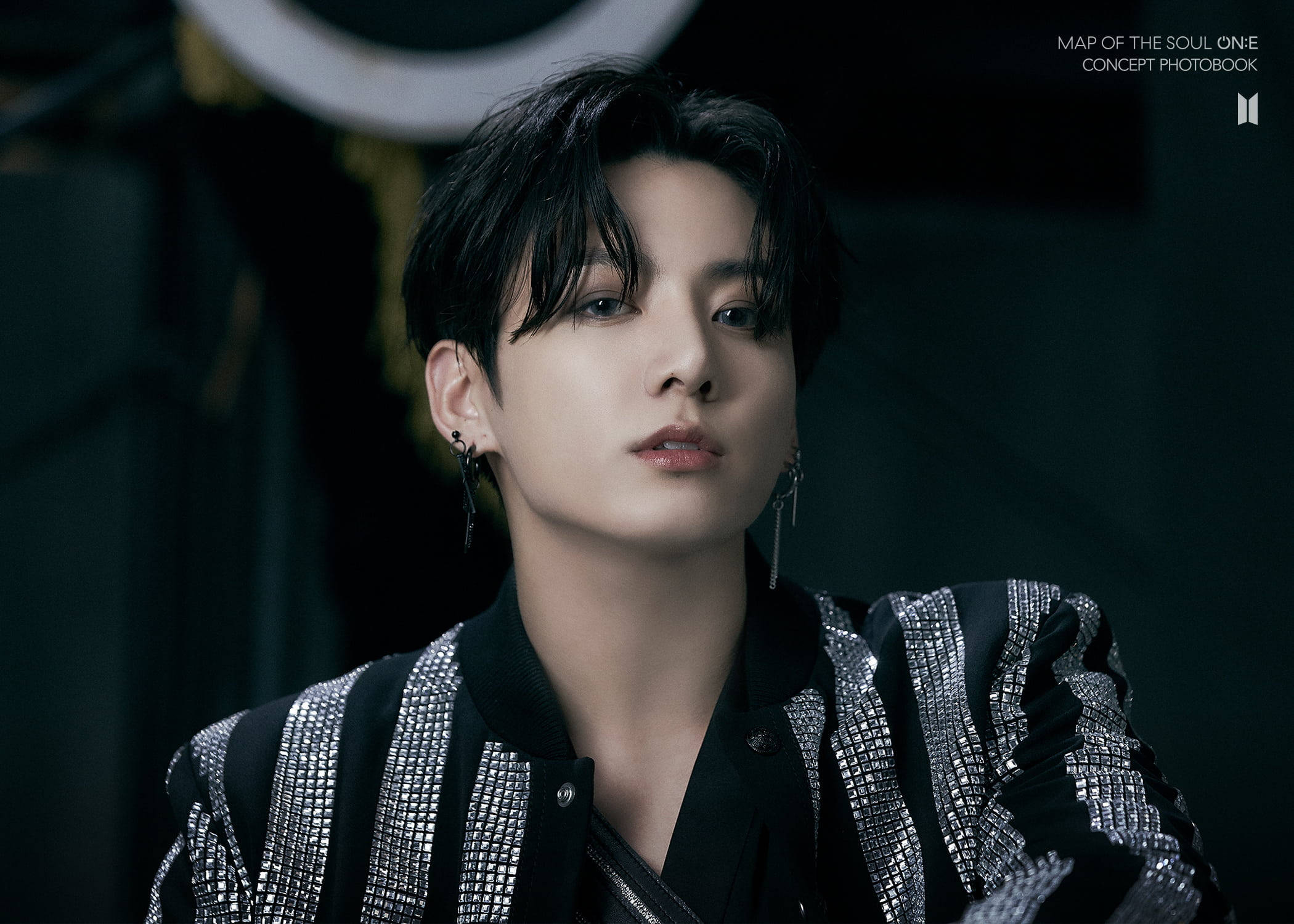 BTS 田柾國 可愛妝容 壁紙 — 一位黑髮戴銀耳環的年輕男子在昏暗的攝影棚中穿著黑色和銀色亮片外套向上凝視。