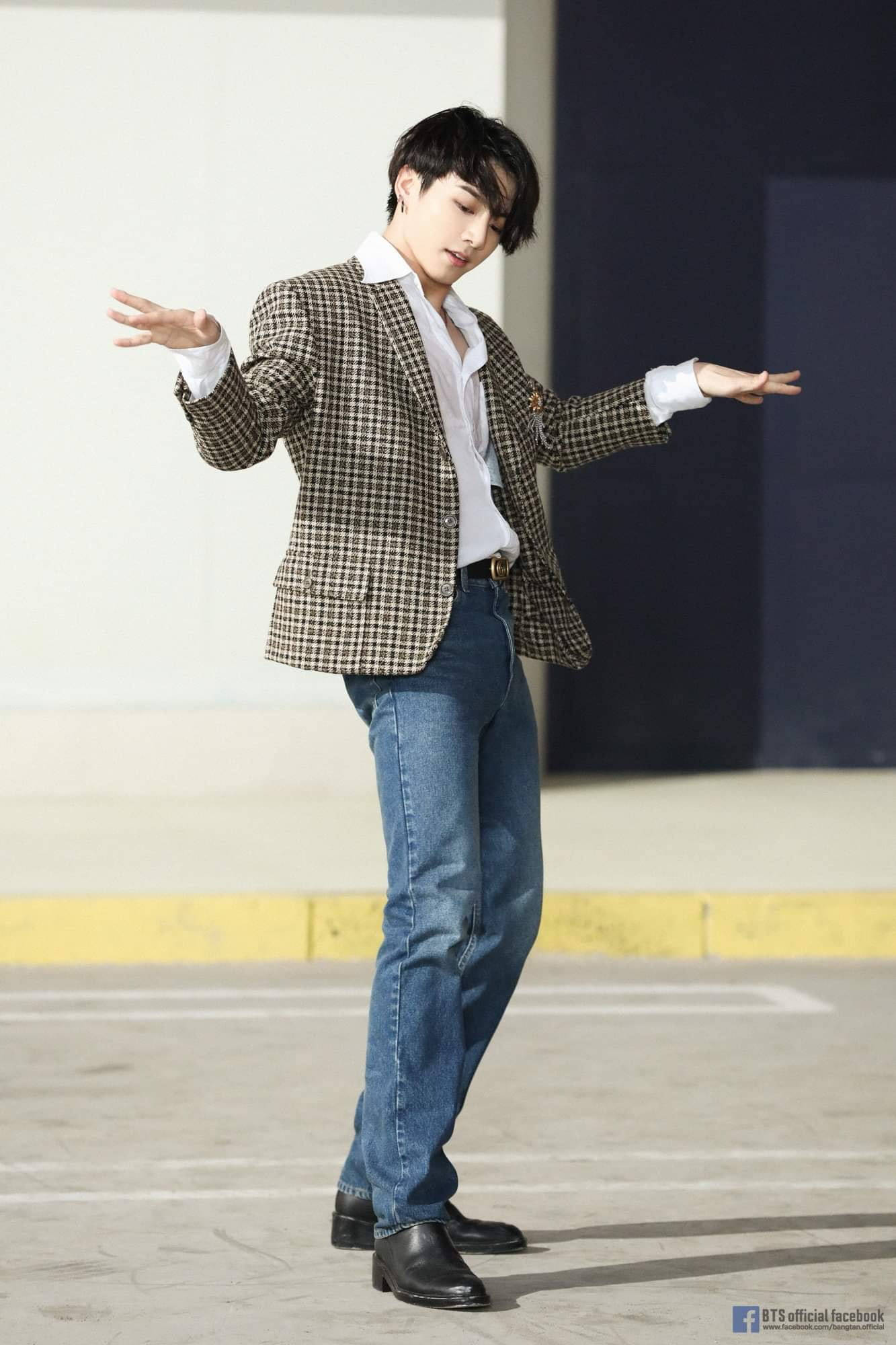 BTS 田柾國 可愛穿搭 壁紙 — 一位身穿格紋西裝外套和牛仔褲的年輕男子，在陽光普照、牆壁對比鮮明的停車場中擺出動態姿勢。