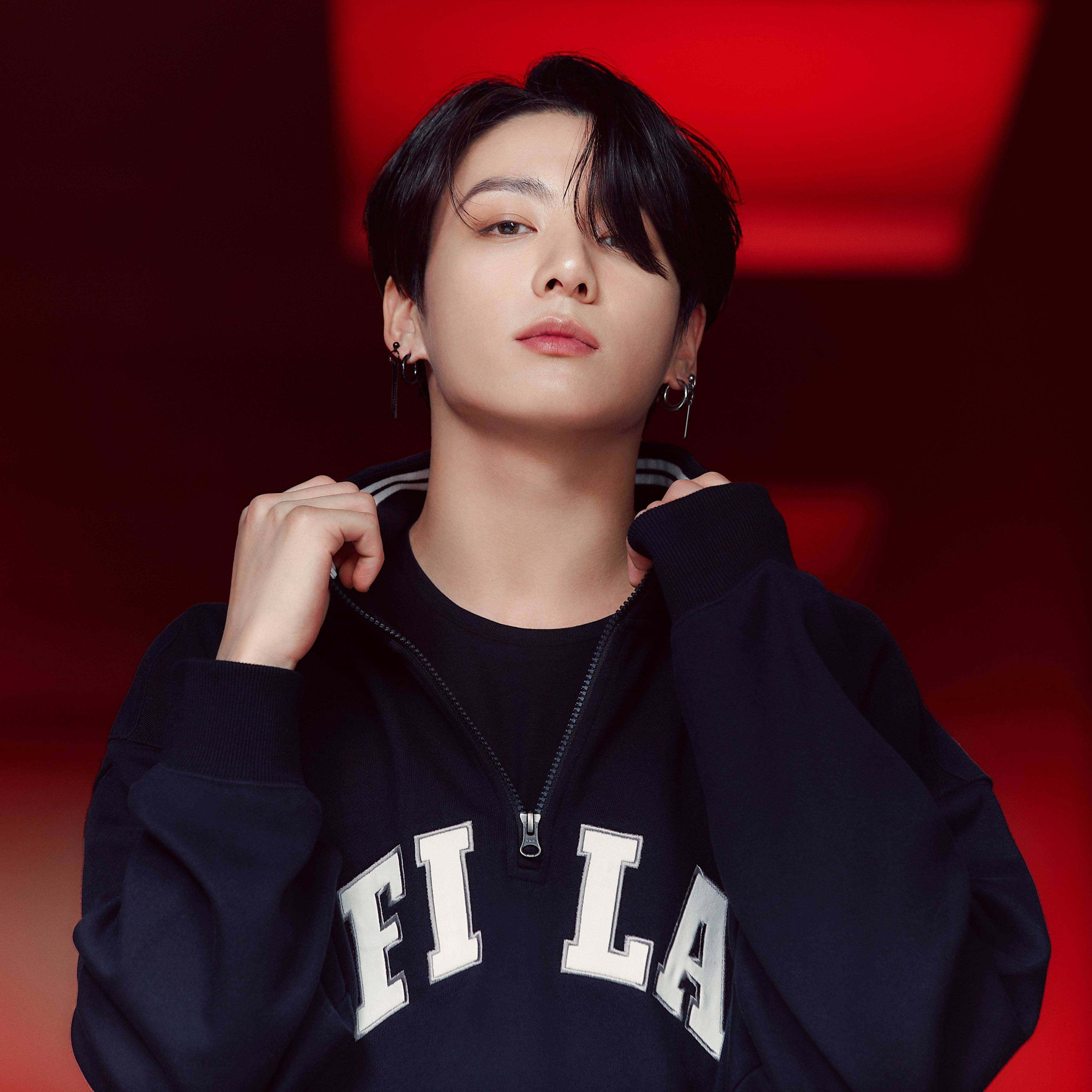 BTS 田柾國 可愛紅色 FILA 壁紙 — 一名深色頭髮、戴著圓環耳環的年輕男子，在鮮紅色的攝影棚背景前拉起黑色Fila運動衫的帽子。