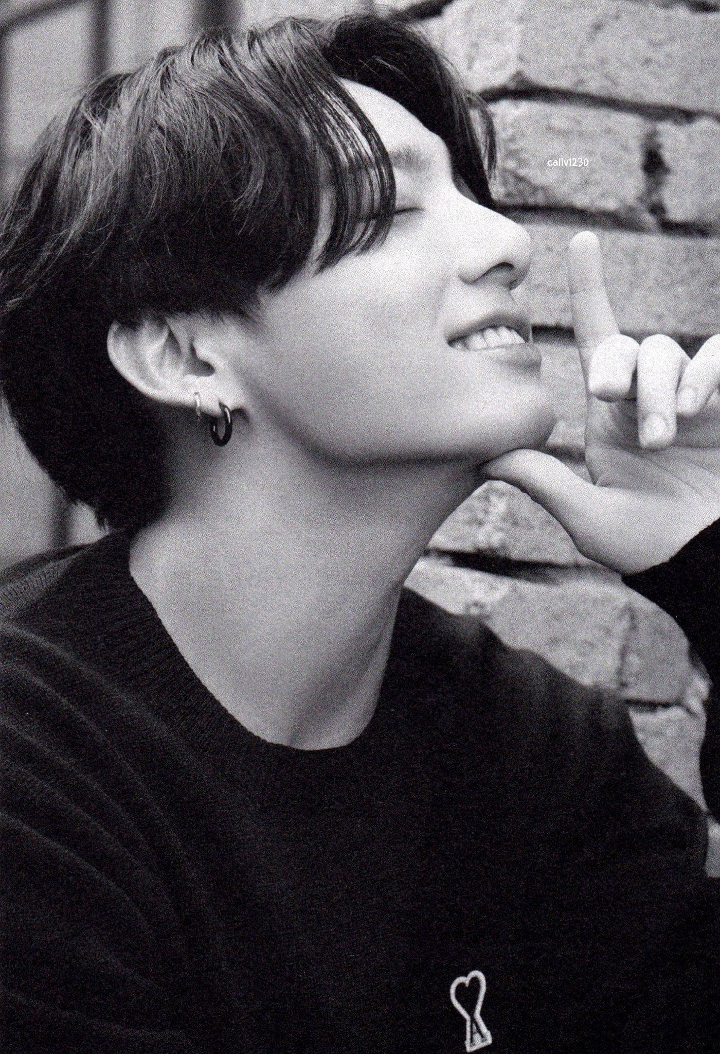 BTS 田柾國 可愛黑白 壁紙 — 一位短黑髮的年輕男子，將下巴擱在手上，靠著磚牆，以黑白風格溫柔微笑。