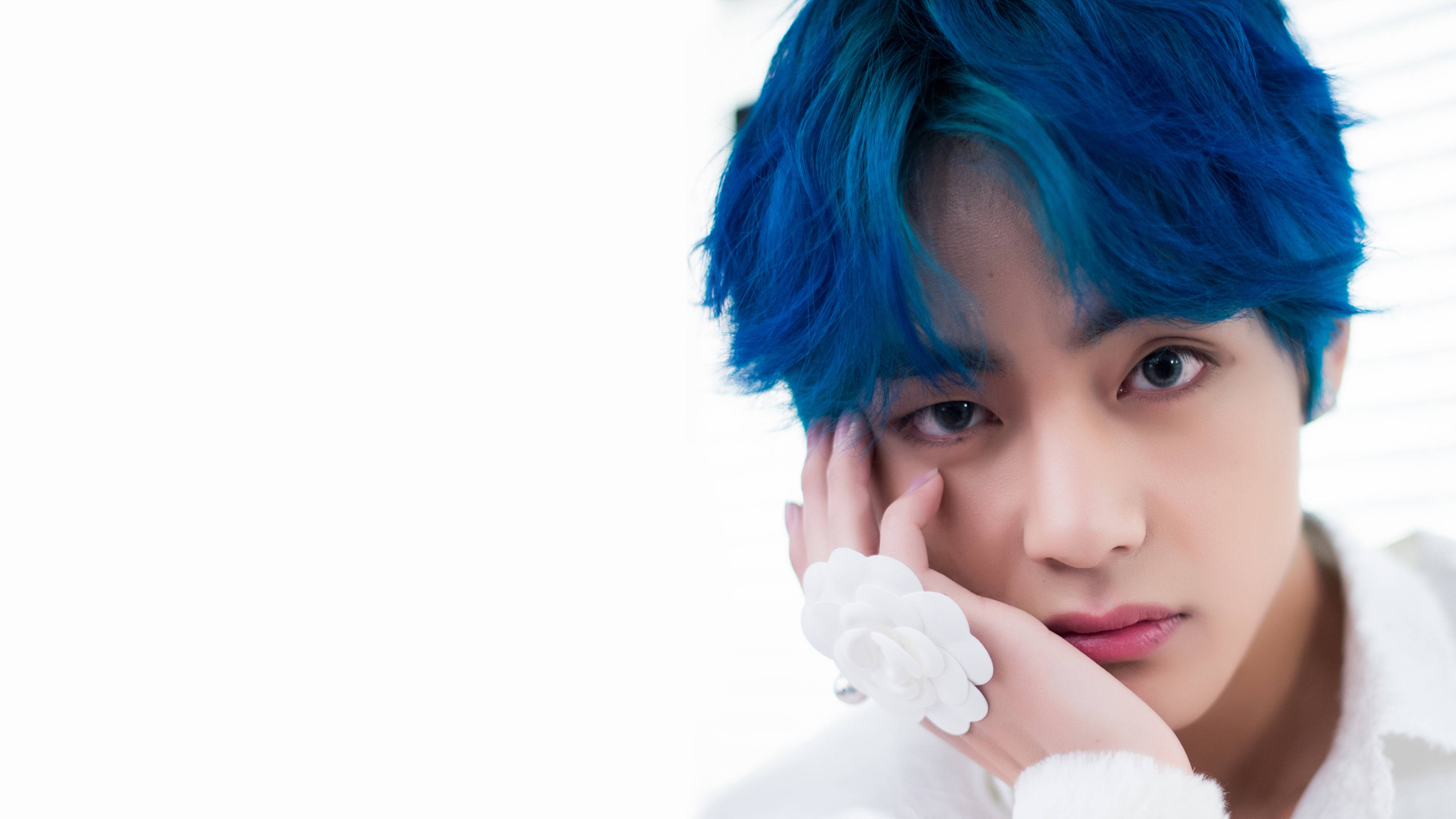 BTS 田柾國 白色花朵戒指 壁紙 — 一位擁有鮮豔藍色頭髮的年輕男子，將下巴擱在戴有白色花卉戒指的手上，背景為明亮的白色。