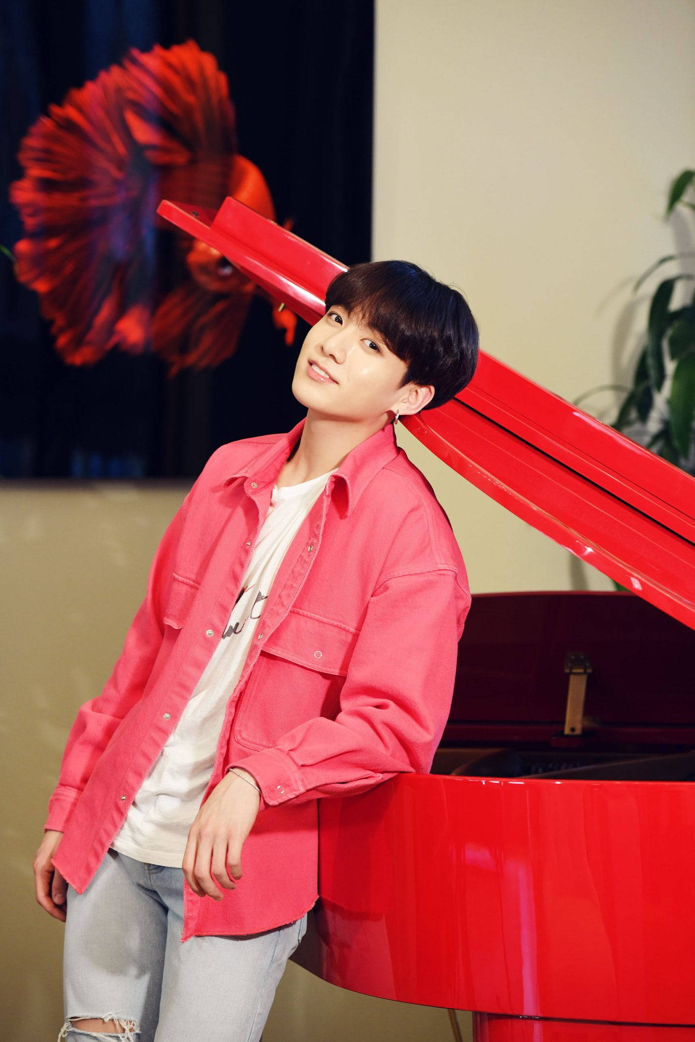 BTS 田柾國可愛微笑 壁紙 — 一名身穿粉紅色襯衫的年輕男子靠在一架光亮的紅色三角鋼琴上，背景中有一條鮮紅色的鬥魚。