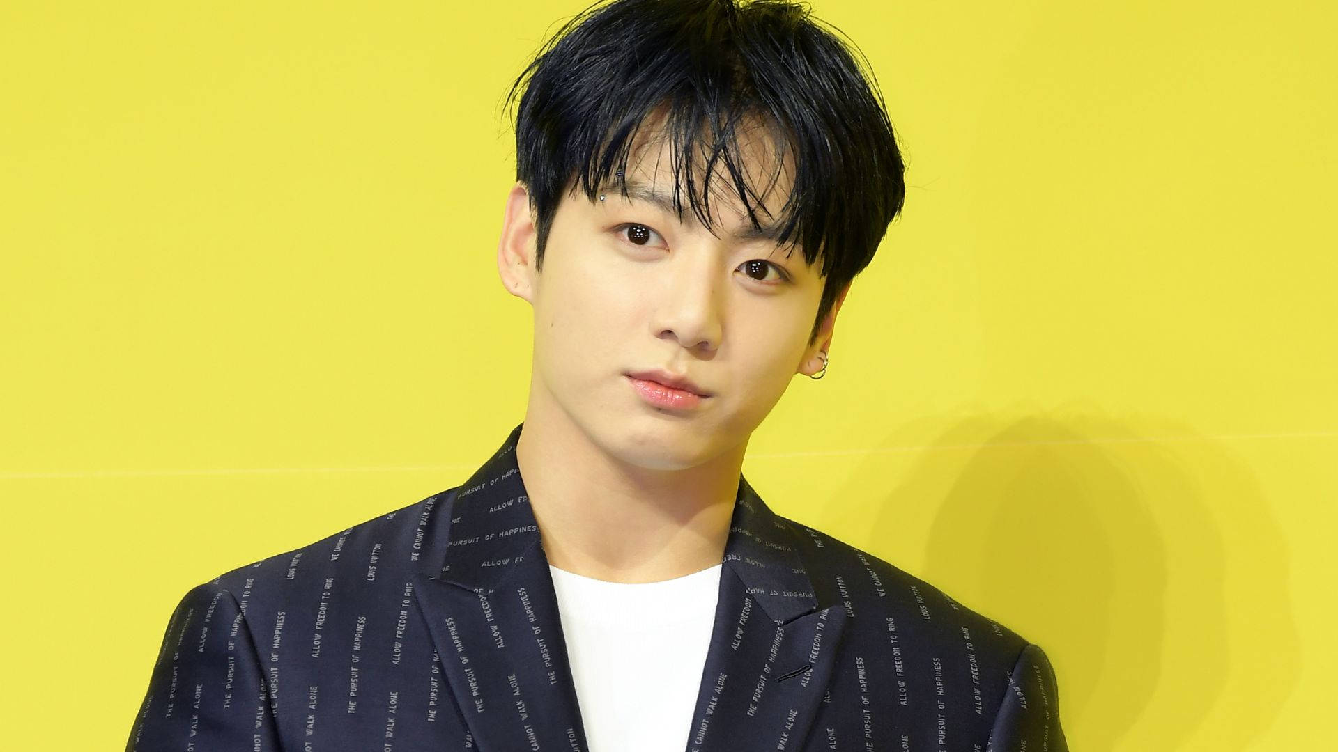 BTS 田柾國黃色背景造型 壁紙 — 一位深色頭髮、戴著低調耳環的年輕男子，在明亮的黃色背景前，穿著一件圖案西裝外套，內搭白色T恤。
