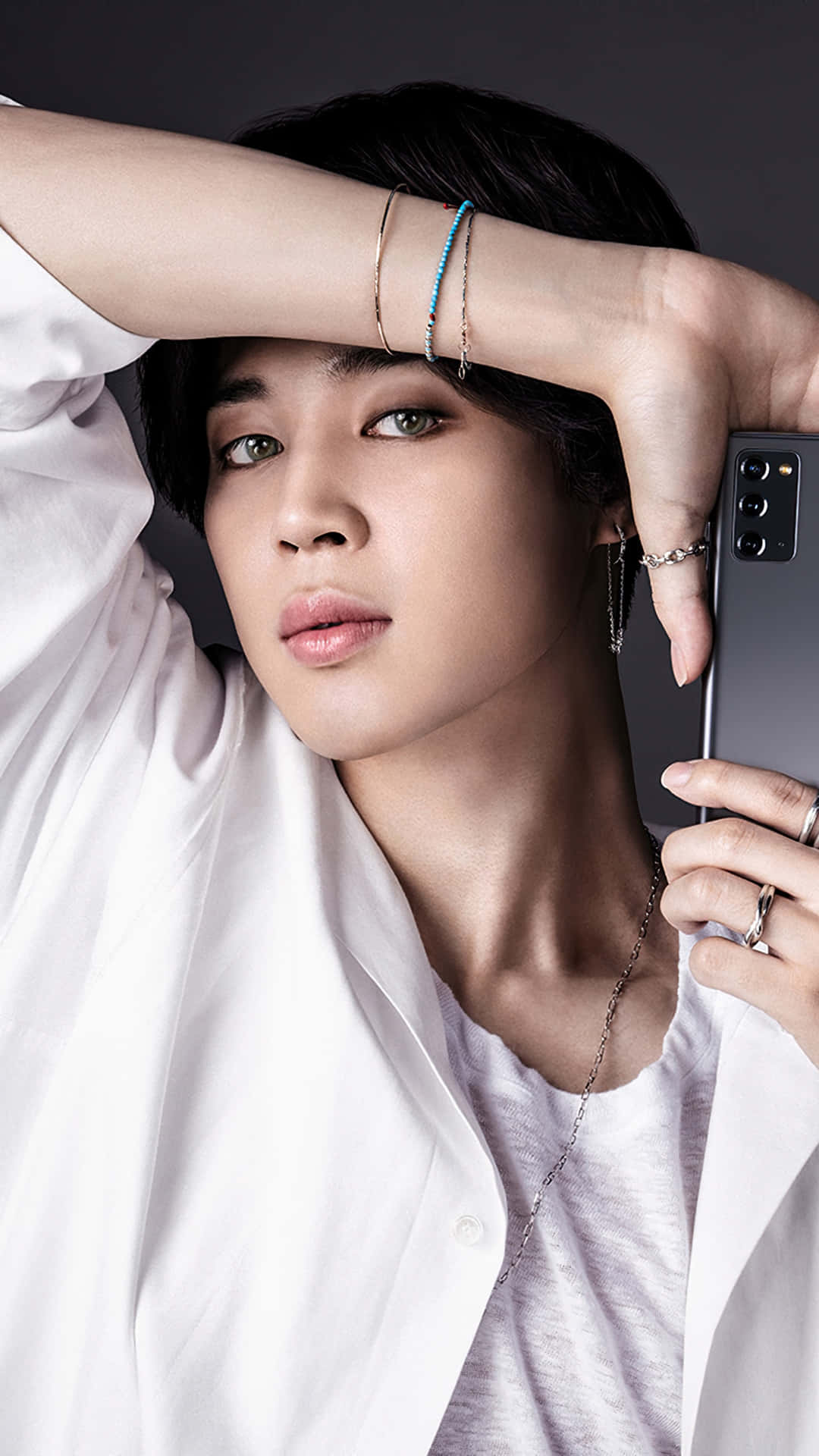 BTS 的多才藝藝人 Jimin 壁紙 — 一位短黑髮綠眼的年輕人，在深色背景前手持智能手機，身穿白襯衫和多條手鐲。