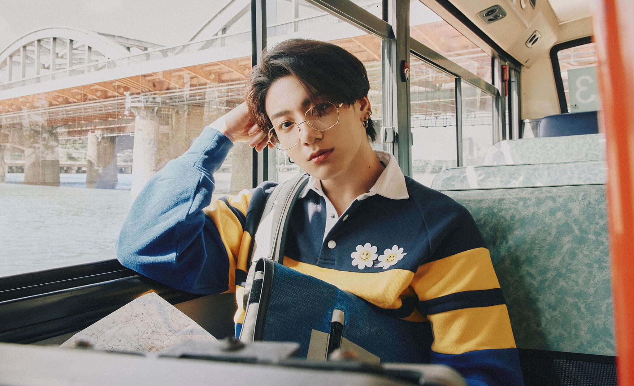 BTS 的才華洋溢 Jungkook 靜態風景照 壁紙 — 一位戴眼鏡的年輕男子靠在巴士窗邊，俯瞰橋樑和水面，身穿藍黃條紋毛衣，帶有花朵補丁。