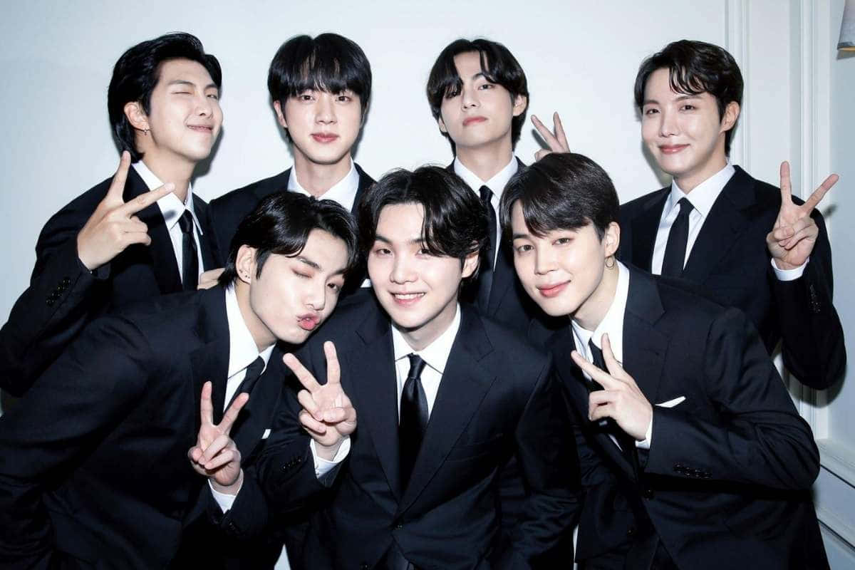 BTS 穿黑色禮服的可愛團體照 壁紙 — 七位時尚的年輕男士身穿黑色西裝，在純白牆前愉快地擺出和平手勢。