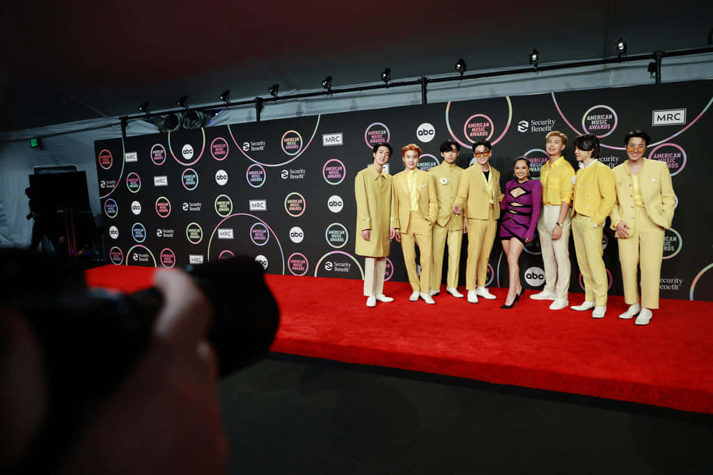 BTS 紅毯照片 壁紙 — 七位穿著時尚黃色西裝的男士和一位穿著紫色服裝的女性在美國音樂獎的紅地毯上自信地擺姿勢。