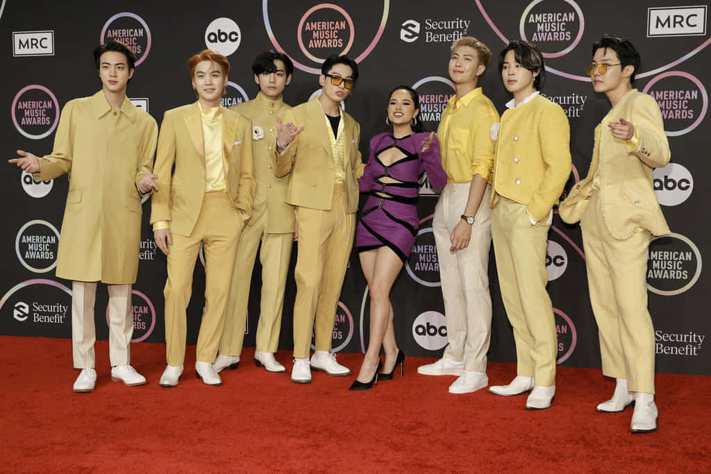 BTS 與歌手Becky G 合照 壁紙 — 一個K-pop組合在美國音樂獎的紅地毯上擺姿勢，有六名身穿黃色西裝的男子和一名身穿紫色連衣裙的女子。