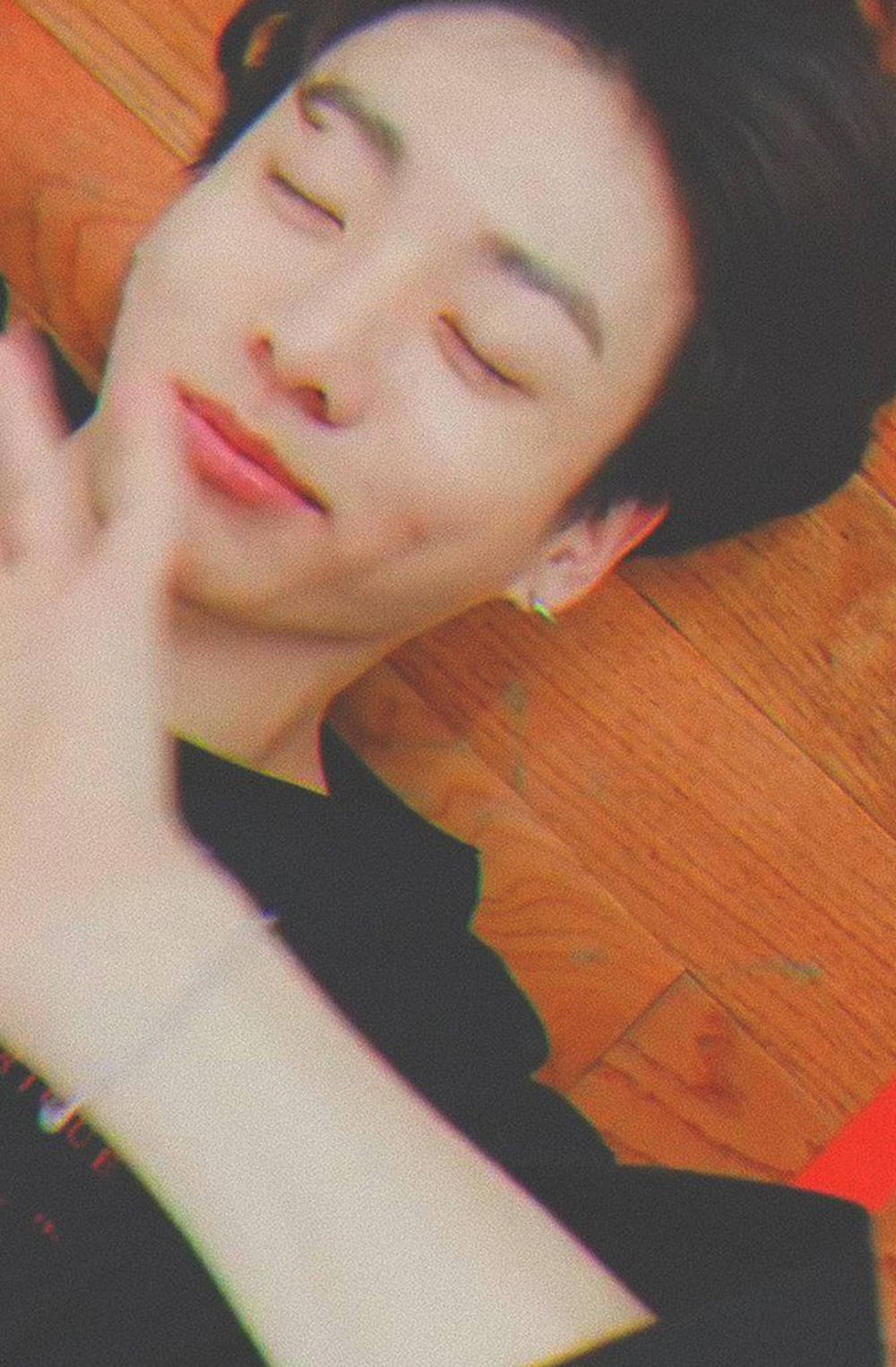 BTS 金俊勉 愛心酒窩特寫 壁紙 — 一名年輕男子閉上眼睛，在溫暖的木地板上平靜地休息，手輕輕舉起靠近臉部。