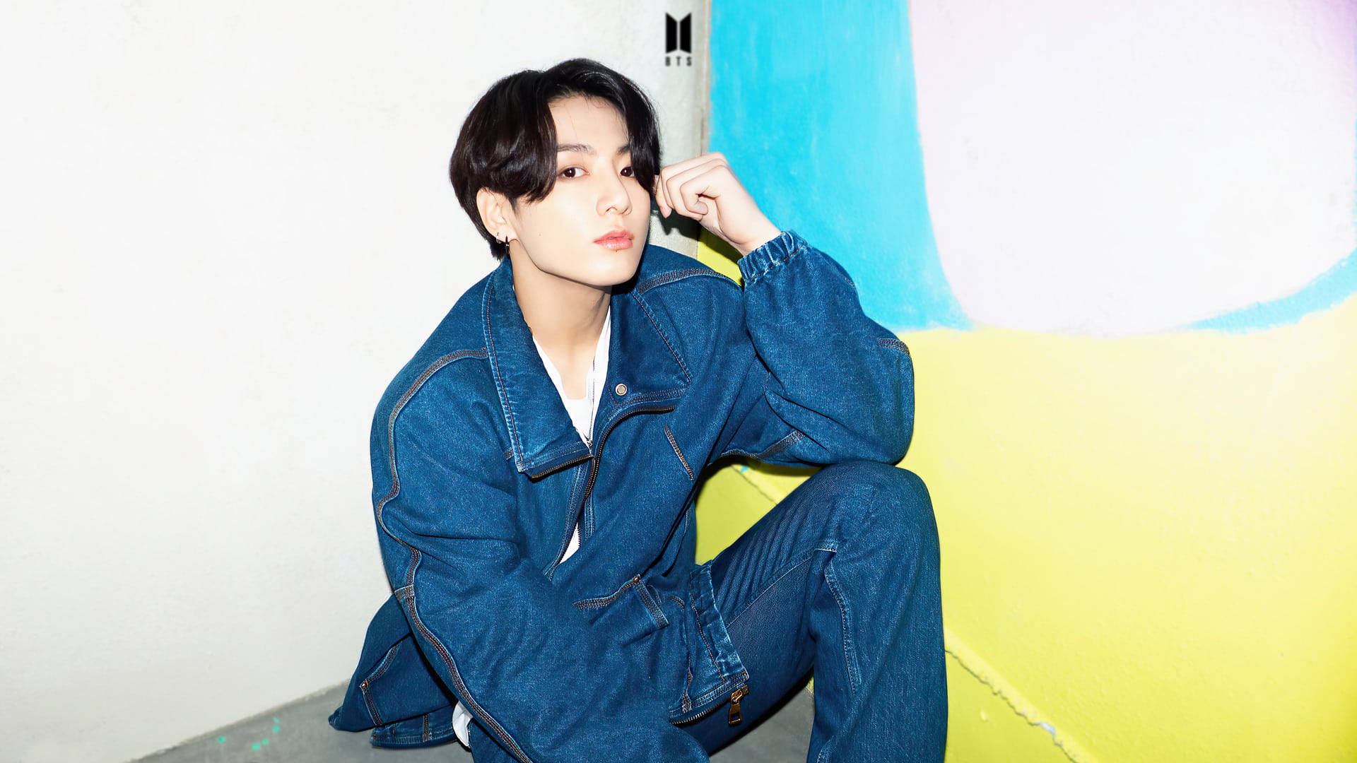 BTS 金俊奎 可愛丹寧造型 壁紙 — 一位身穿牛仔服的年輕男子隨意地坐在一面色彩繽紛、帶有醒目藍色、黃色和白色區塊的牆前。
