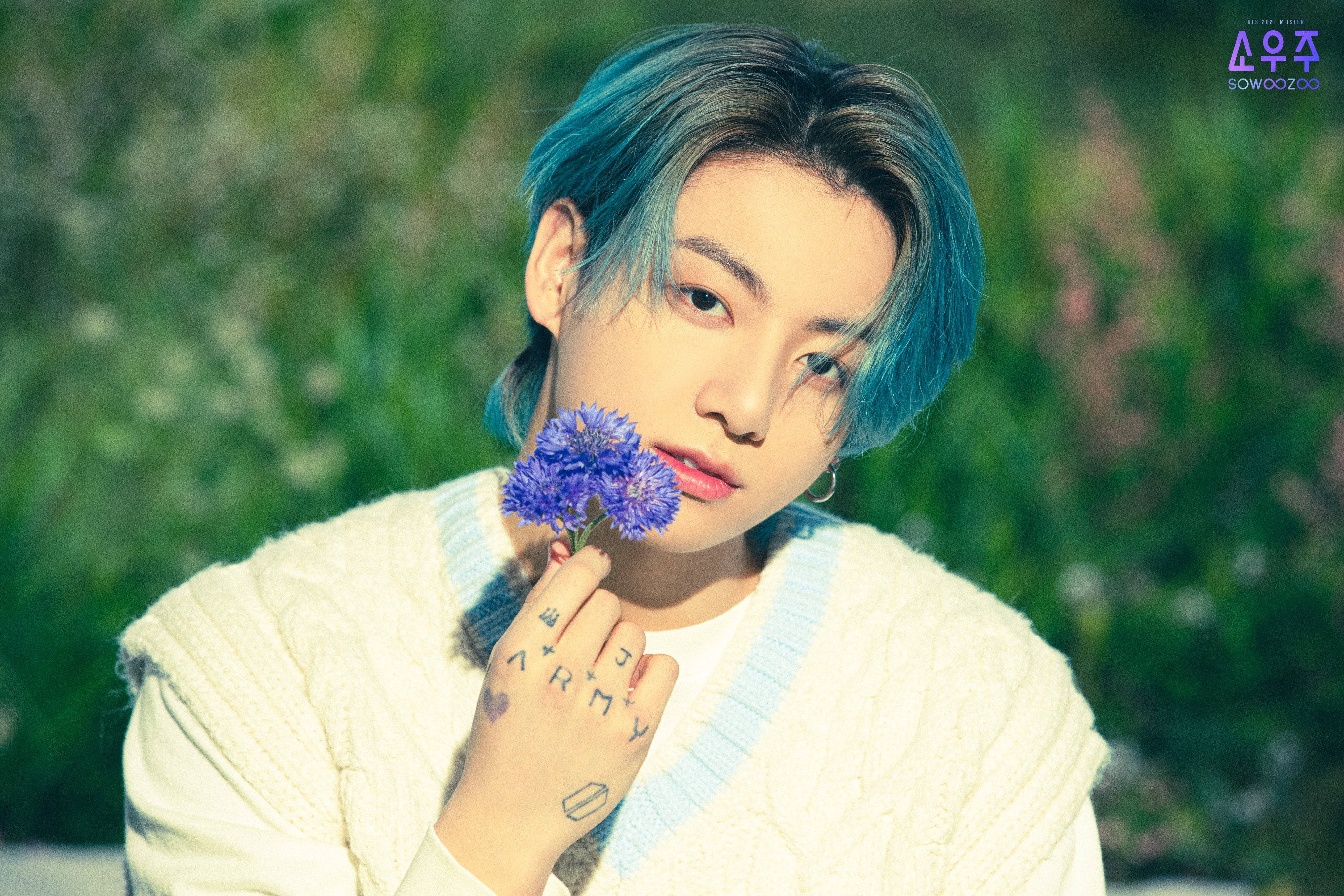 BTS 金俊奎 可愛藍髮造型 壁紙 — 一個藍色頭髮的年輕人，在柔和的綠色田野背景下，將一朵紫色花朵靠近臉龐。