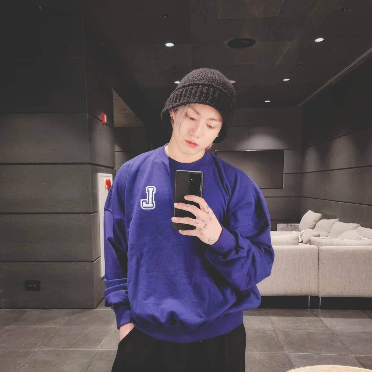 BTS 金俊奎 可愛鏡前自拍 壁紙 — 一個人穿著藍色衛衣和黑色冷帽，在一個灰色牆壁和米色梳化的現代簡約房間裡對著鏡子自拍。