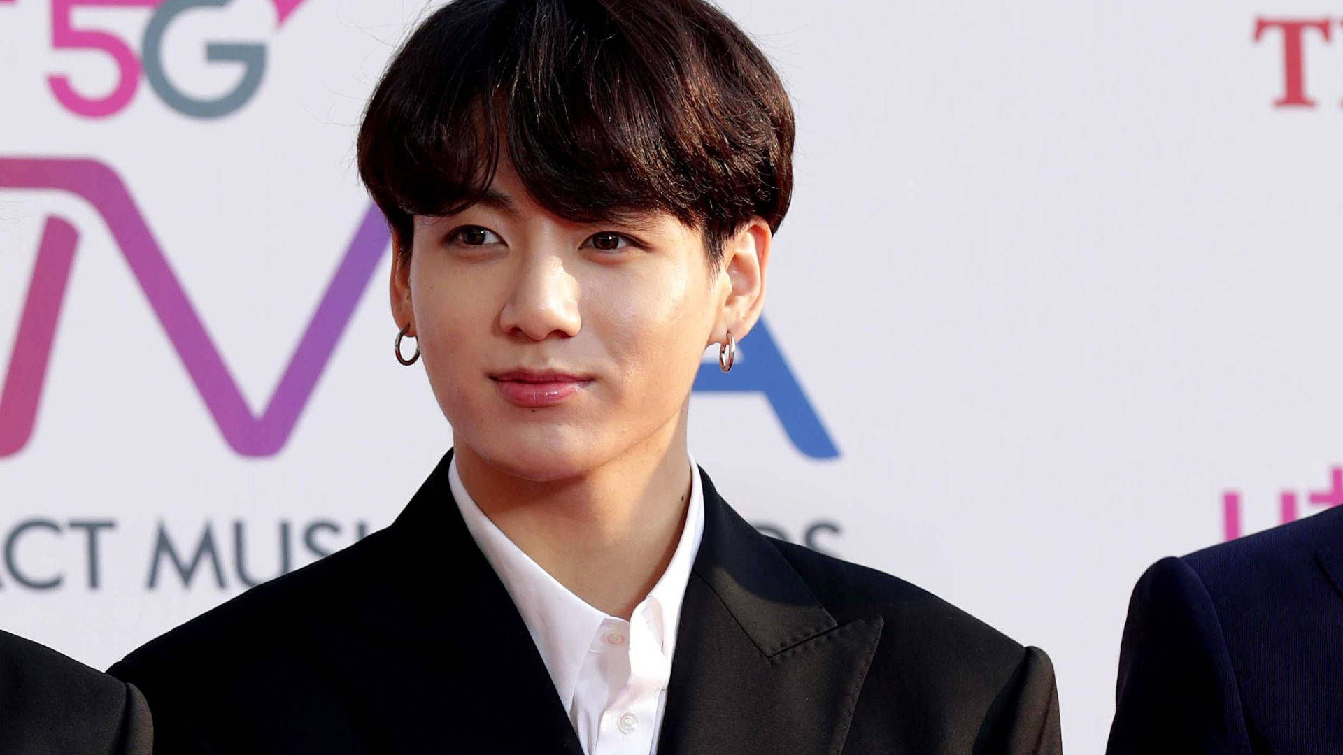 BTS 金俊奎 紅毯 壁紙 — 一位深色頭髮、戴著圈狀耳環的年輕男子，在音樂頒獎活動中穿著黑色西裝和白色襯衫。