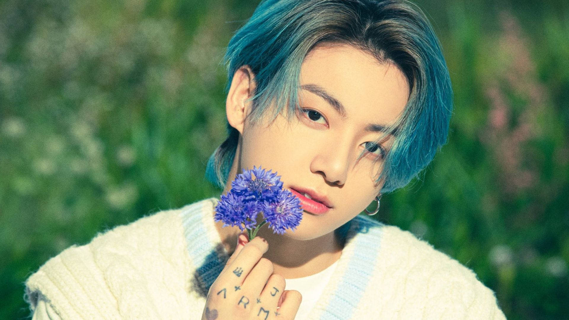 BTS 金俊奎 紫色花朵 壁紙 — 一個藍色頭髮的年輕人，在柔和的綠色田野背景下，將一朵紫色花朵靠近臉龐。