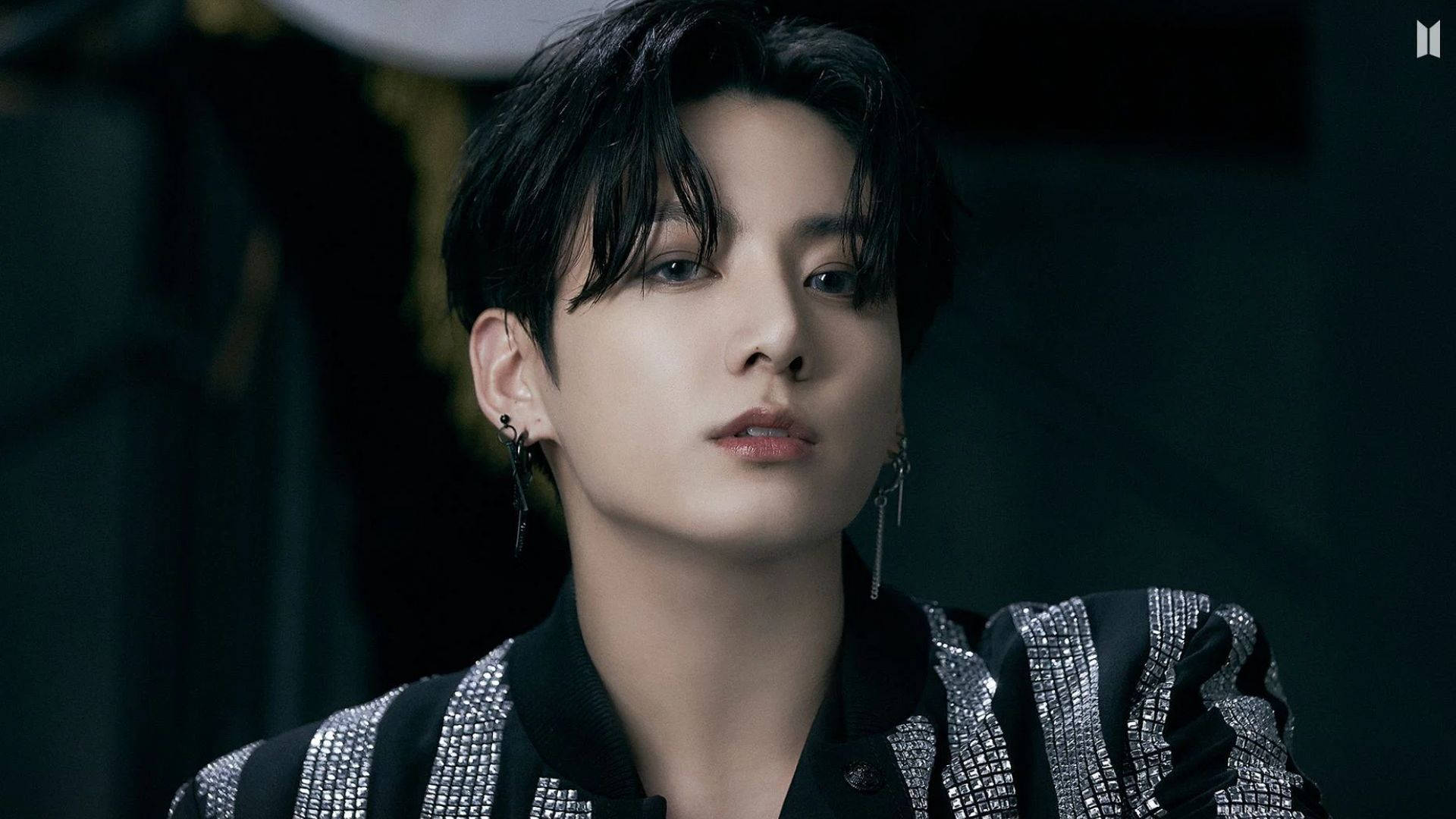 BTS 金俊奎 高級風 壁紙 — 一個年輕黑髮男子戴著垂墜耳環，身穿黑色和銀色裝飾外套，在昏暗背景下的特寫肖像。