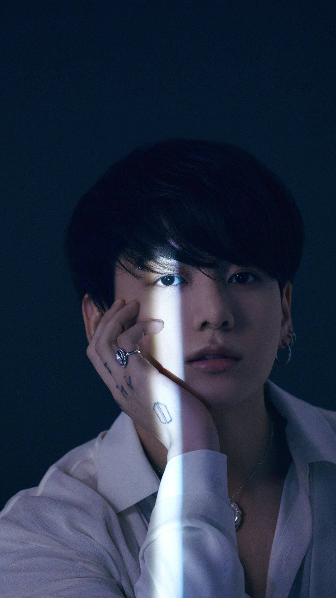 BTS 金俊奎 黑暗中 壁紙 — 一位深色頭髮的年輕男子將臉靠在手上，被一道垂直的光束照亮，背景為深藍色。