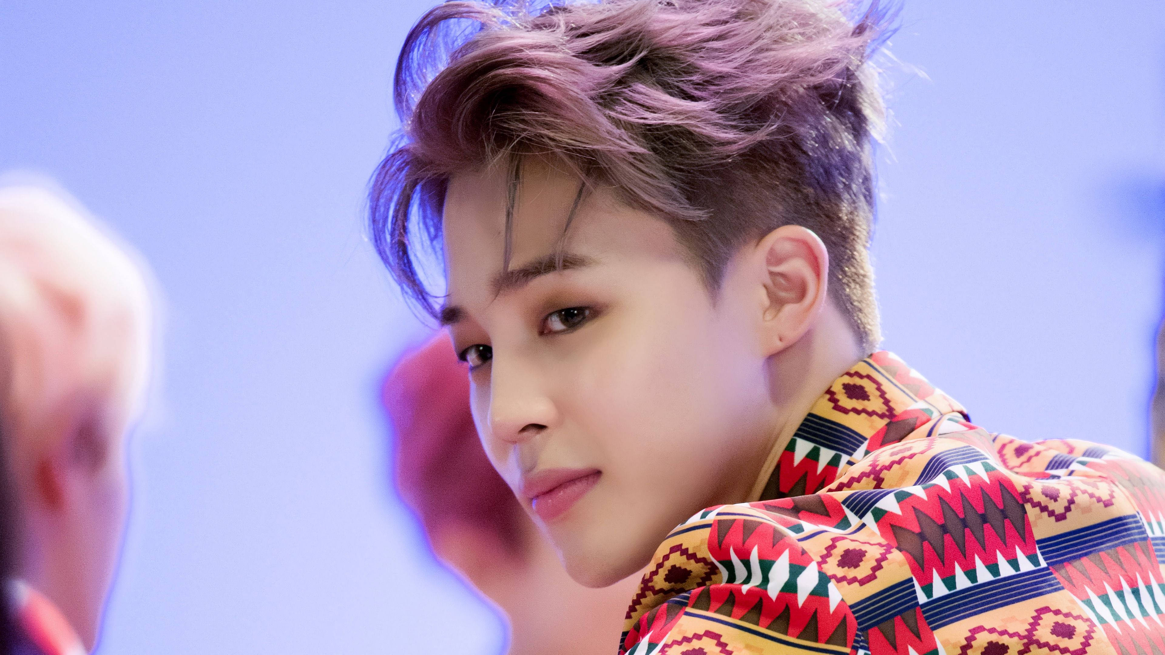 BTS 金珍洙《IDOL》MV造型 壁紙 — 一位年輕男子，帶有紫色調的頭髮和色彩繽紛的幾何圖案襯衫，在柔和的藍色背景下若有所思地凝視。
