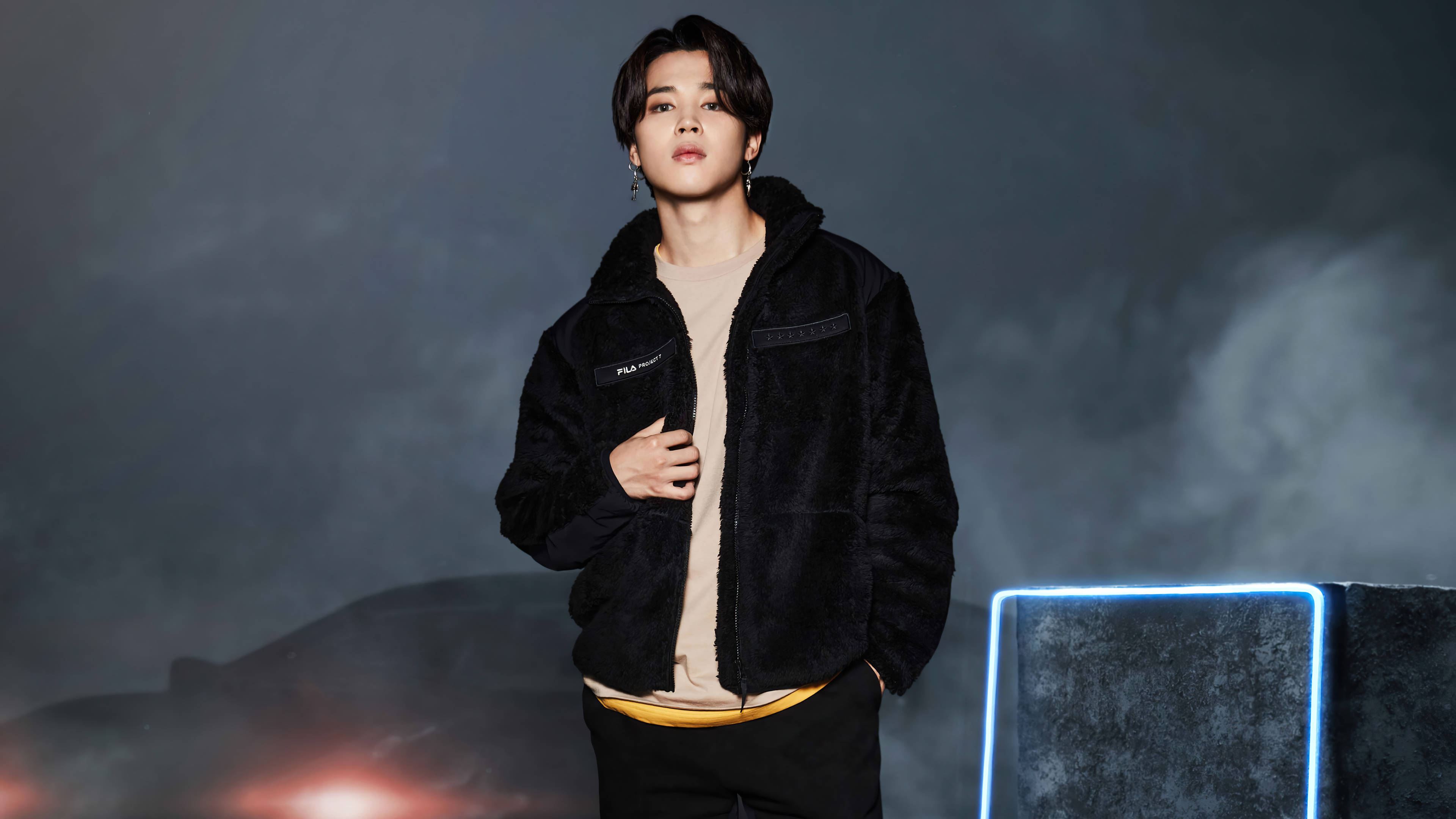 BTS 金珍洙為Fila代言 壁紙 — 一個穿著黑色毛絨外套的年輕男子自信地站在煙霧瀰漫的黑暗背景前，帶有發光的藍色霓虹燈點綴。
