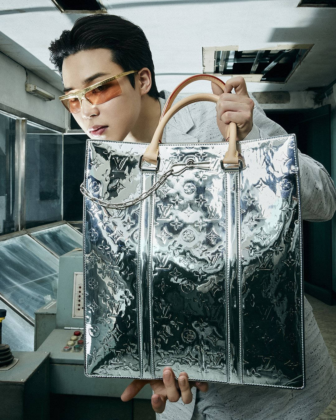 BTS 金珍洙為Louis Vuitton代言 壁紙 — 一個戴著金色邊框太陽眼鏡的男人，在復古工業環境中拿著一個帶有壓花字母組合的大型銀色路易威登手袋。