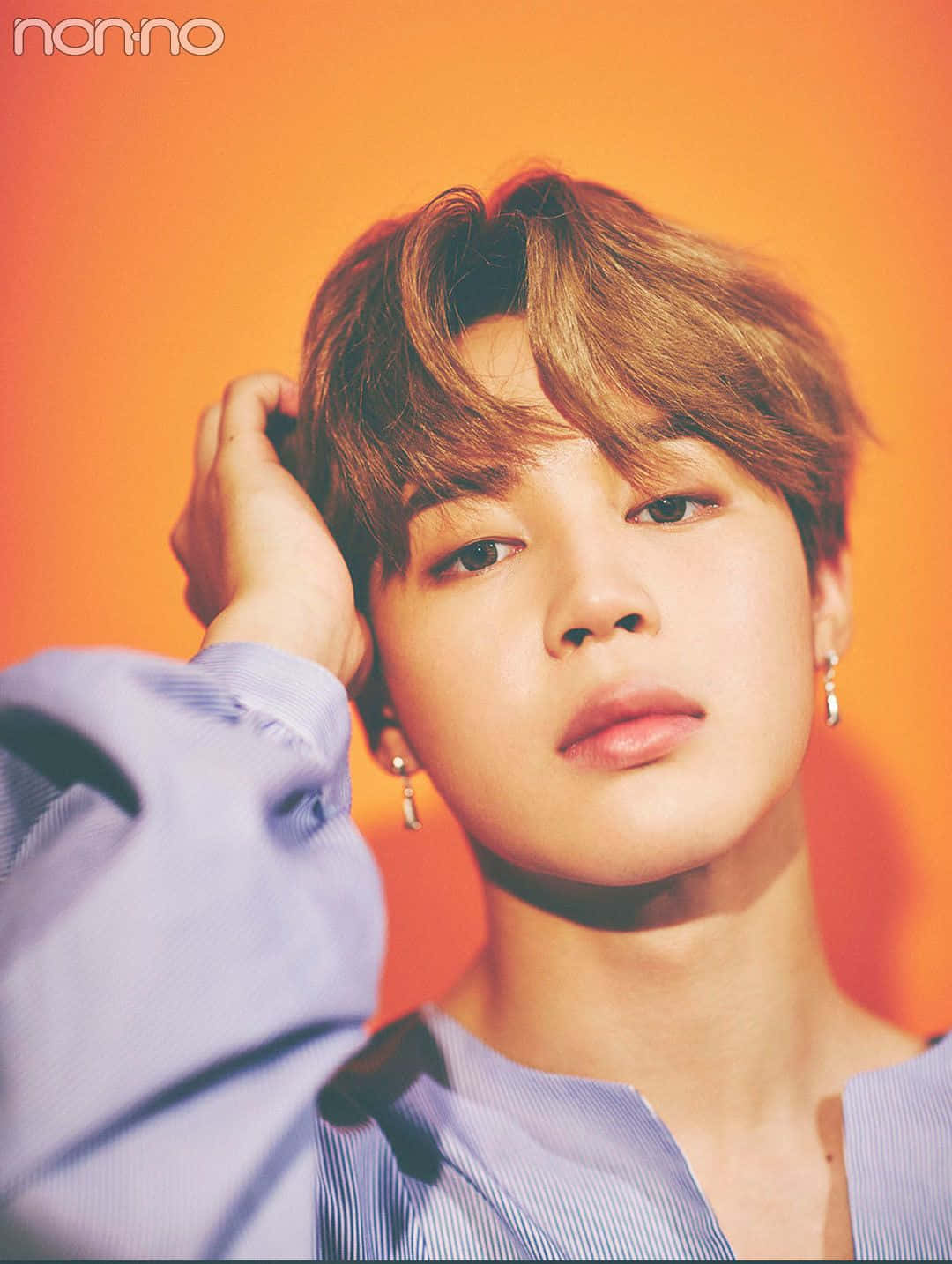 BTS 領唱者 Jimin，鼓舞人心的音樂傳奇 壁紙 — 一位年輕男子，一頭凌亂的棕色頭髮，戴著銀色耳環，在溫暖的橙色背景下柔和地凝視，同時用一隻手觸摸自己的頭。
