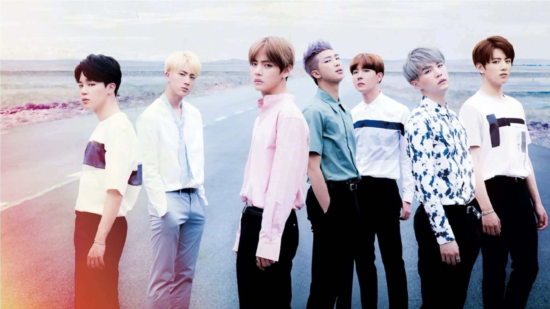 BTS加入蘋果陣營 壁紙 — 七位時尚年輕男子身穿彩色襯衫，在柔和的天空下自信地站在沙漠公路旁。