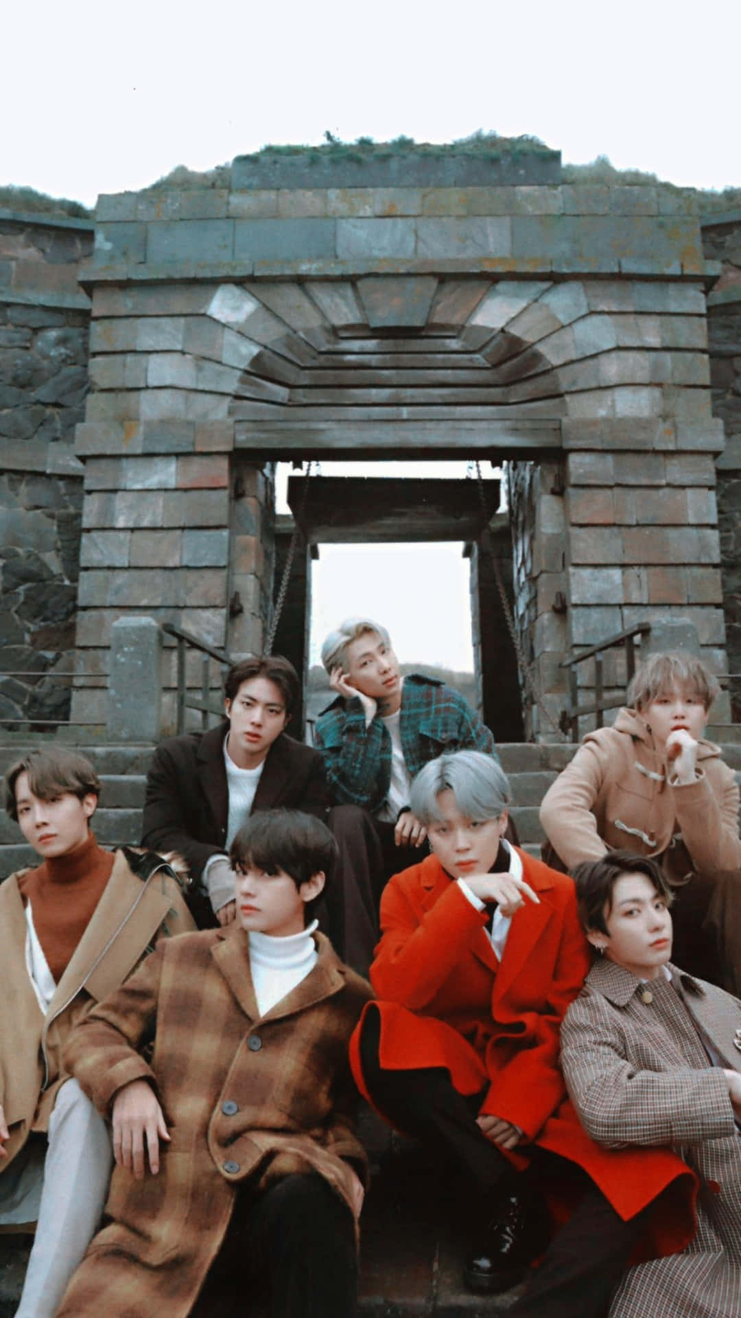 BTS坐石階拍攝 壁紙 — 七位時尚的年輕男子穿著秋季外套，在風化的石拱門下的石階上擺姿勢。