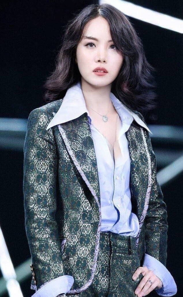 身穿圖案綠色西裝外套和藍色襯衫的女士，在明亮燈光下站在舞台上。