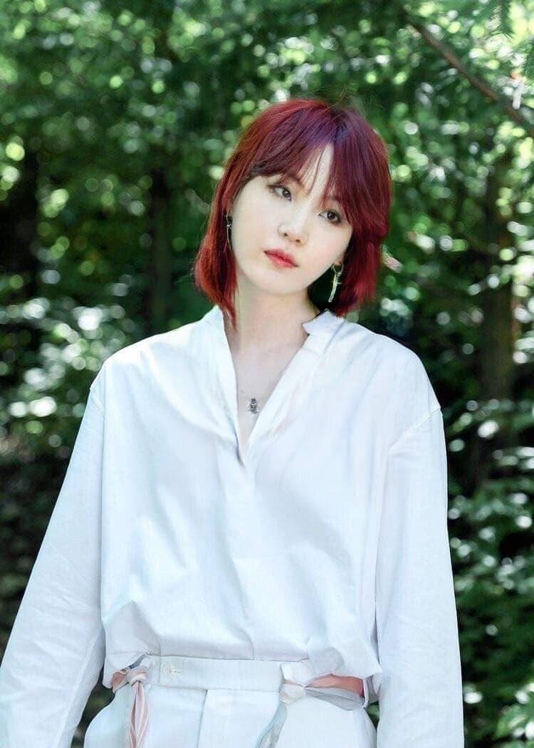 紅髮女性身穿白色服裝，站在陽光普照的森林中。