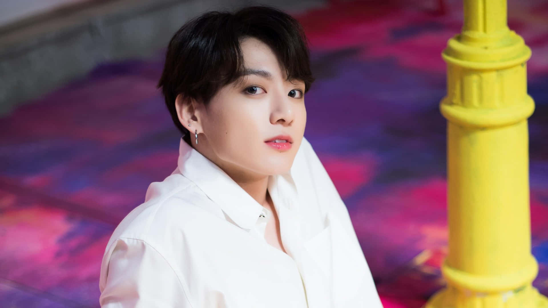 BTS成員Jungkook在攝影棚展露微笑 壁紙 — 一位身穿白恤衫的年輕男子在鮮黃色柱子旁邊，溫柔地凝視著色彩繽紛的紫粉紅色地板。