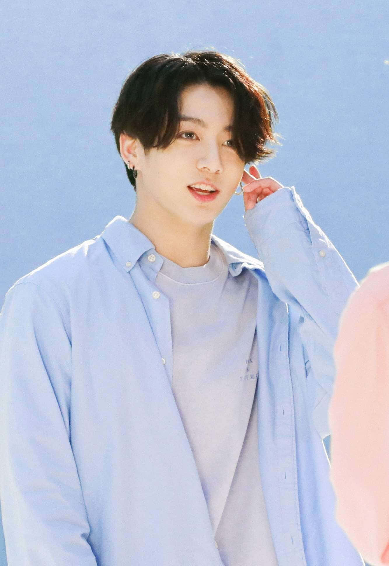 BTS成員Jungkook展現迷人姿態 壁紙 — 一位黑髮戴耳環的年輕男子在清澈的藍天下輕輕微笑，一邊整理他的淺藍色鈕扣襯衫。