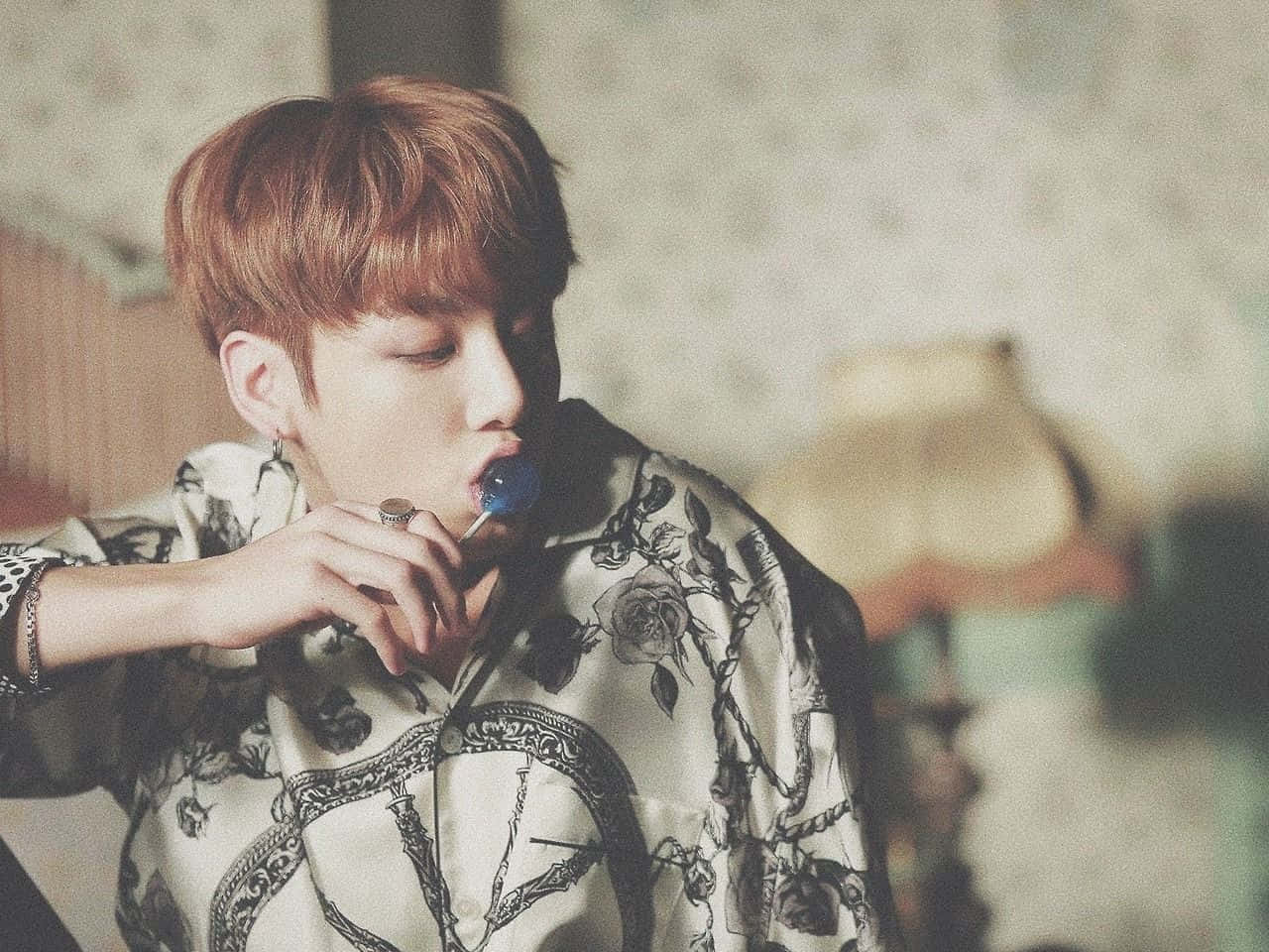 BTS成員Jungkook展露自信笑容 壁紙 — 一位紅髮年輕男子穿著花卉圖案襯衫，在柔和光線的房間裡舔著藍色棒棒糖。
