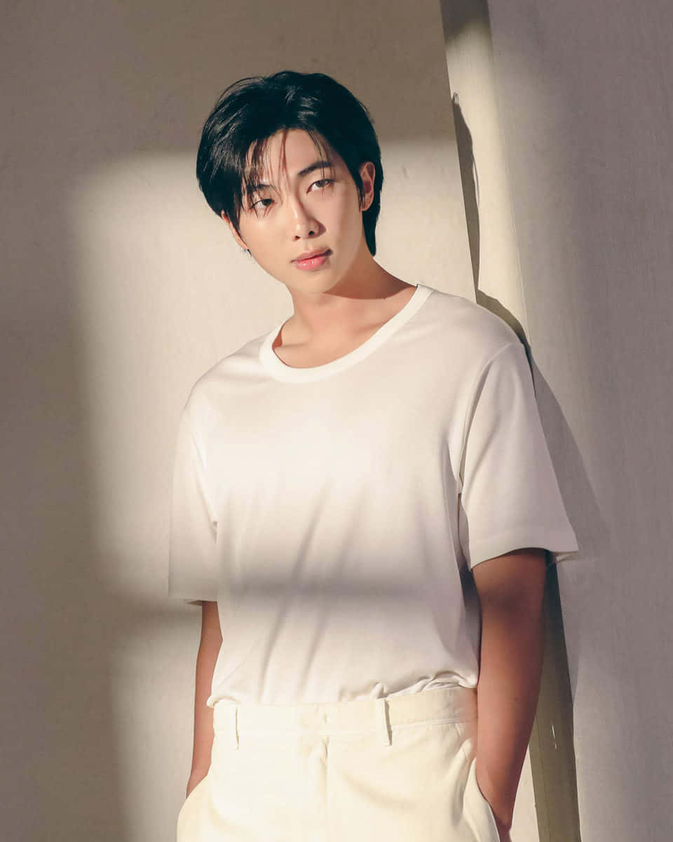 BTS成員RM的經典造型 壁紙 — 一位身穿白色T恤和褲子的年輕男子靠在一面牆上，牆上灑滿柔和自然光，投下輕柔陰影。