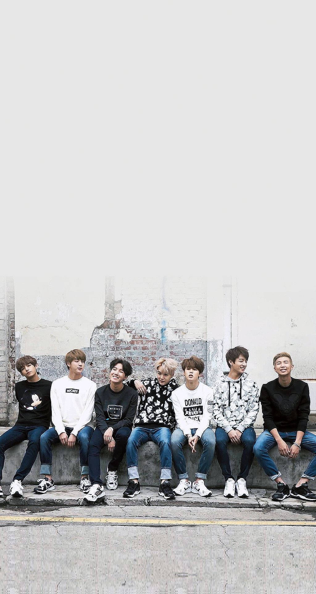 BTS手機螢幕模糊背景展示 壁紙 — 七位身穿休閒街頭服的年輕男性，隨意地坐在風化的混凝土邊緣上，背景是褪色的磚牆。