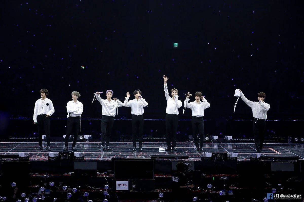 七位K-pop偶像在燈光璀璨的舞台上表演，背景是星空，粉絲們在歡呼。