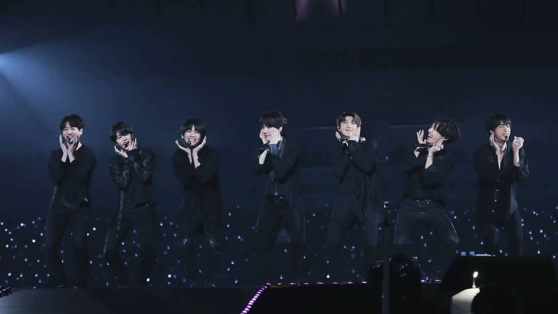 七位男性K-pop表演者在黑暗舞台上，伴隨藍色燈光和閃爍地板演唱。