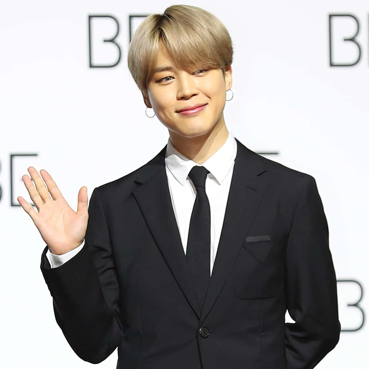 BTS的Jimin以獨特風格展現魅力 壁紙 — 一位微笑的金髮男士穿著黑色西裝，在白色品牌背景前揮手。