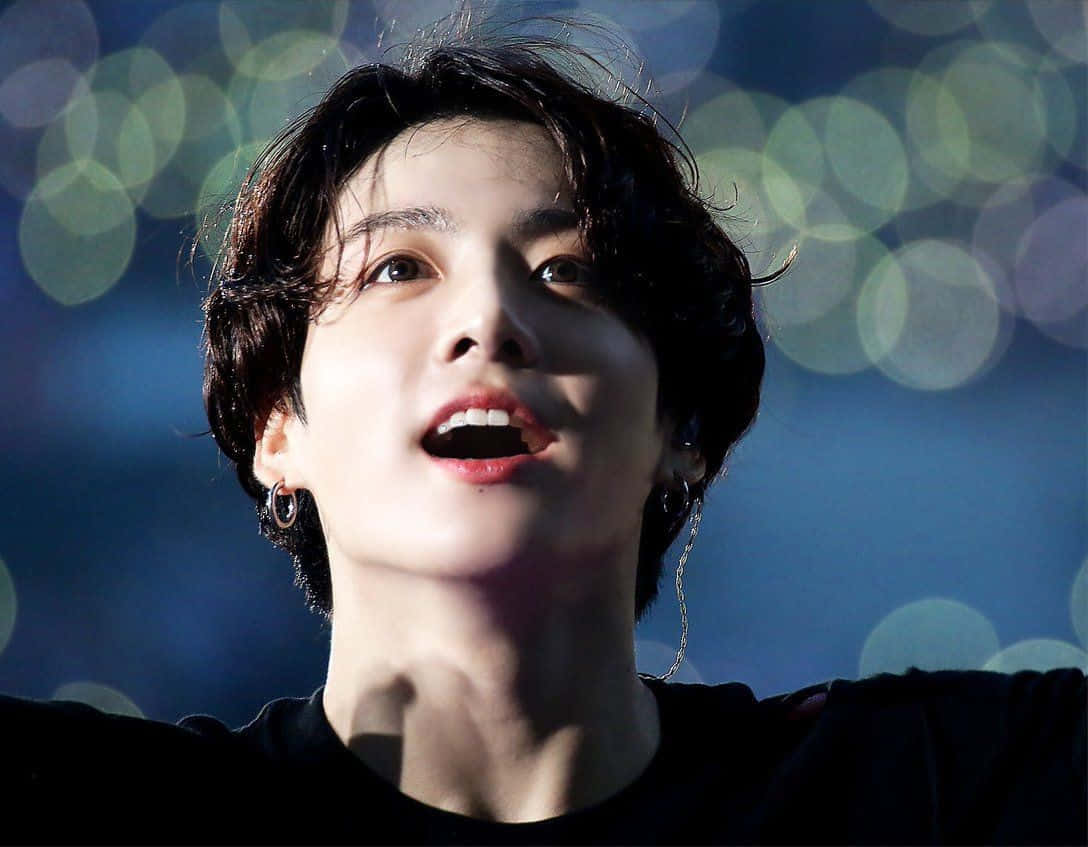 BTS的Jungkook，魅力男神 壁紙 — 一位深色頭髮、戴著耳環的年輕男子，在藍色散景背景下張開嘴巴向上凝視。