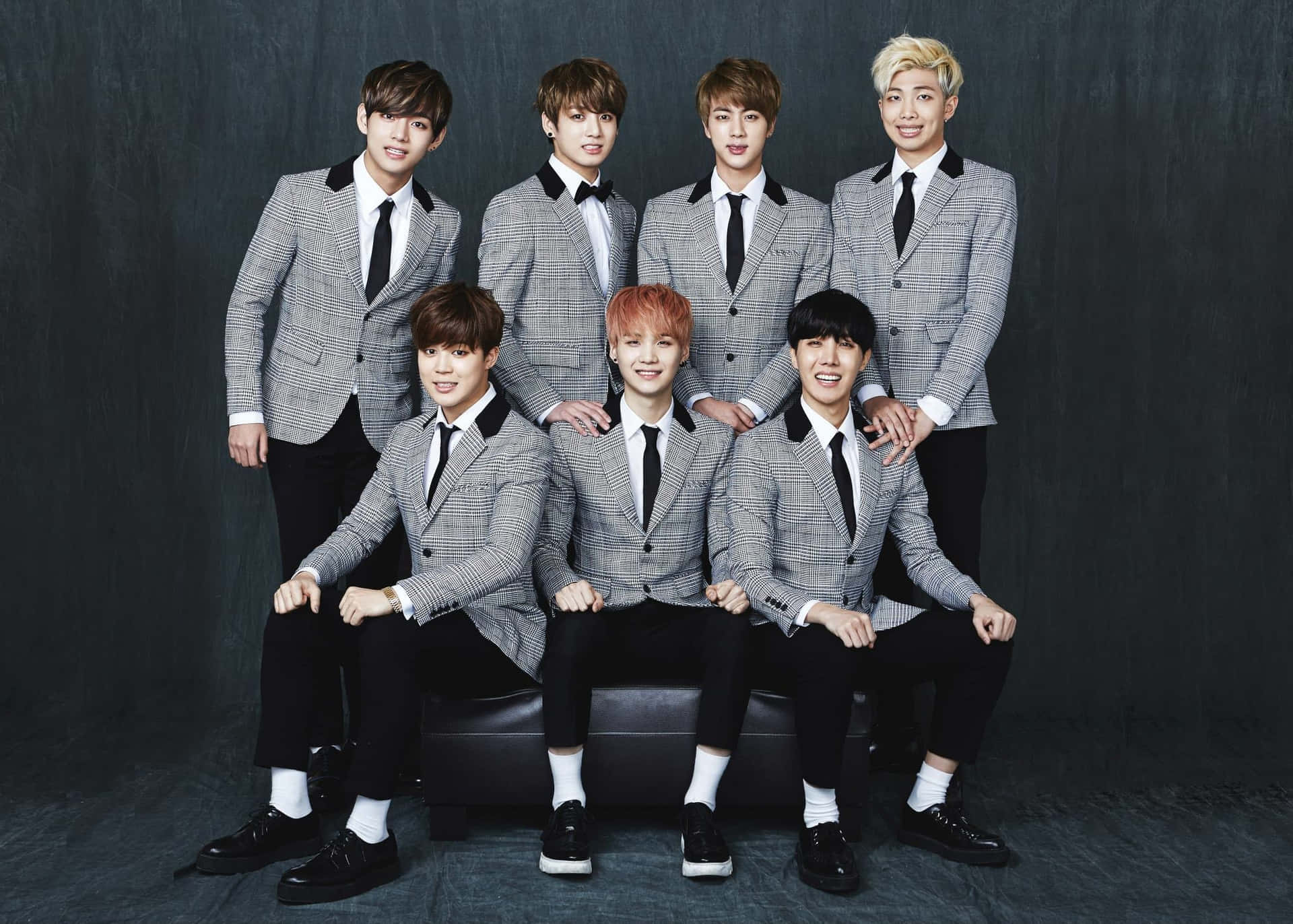 BTS穿格紋西裝拍攝 壁紙 — 七位年輕男士身穿相配的灰色格紋西裝，自信地坐在黑色長椅上，背景是深色紋理背景。