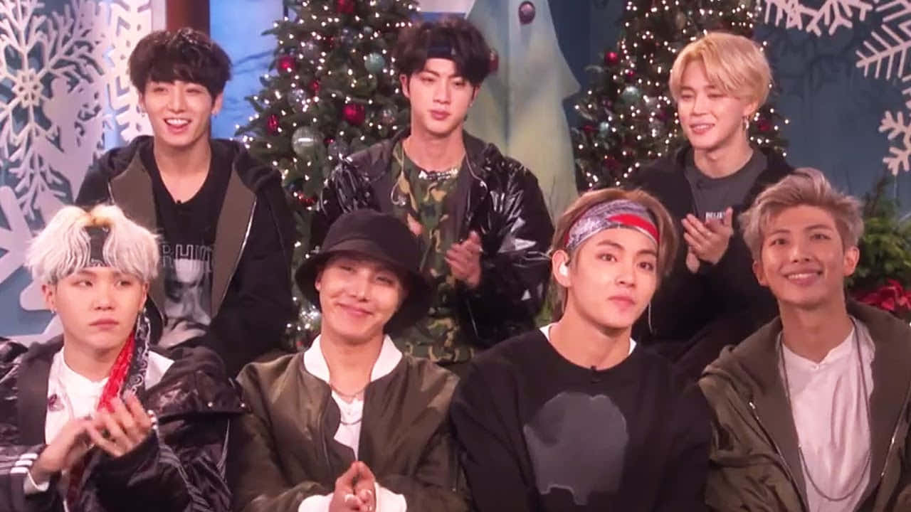 BTS聖誕特別訪問照片 壁紙 — 七位微笑的年輕男士穿著時尚的冬季服裝，在裝飾好的聖誕樹和雪花背景前開心地擺姿勢。