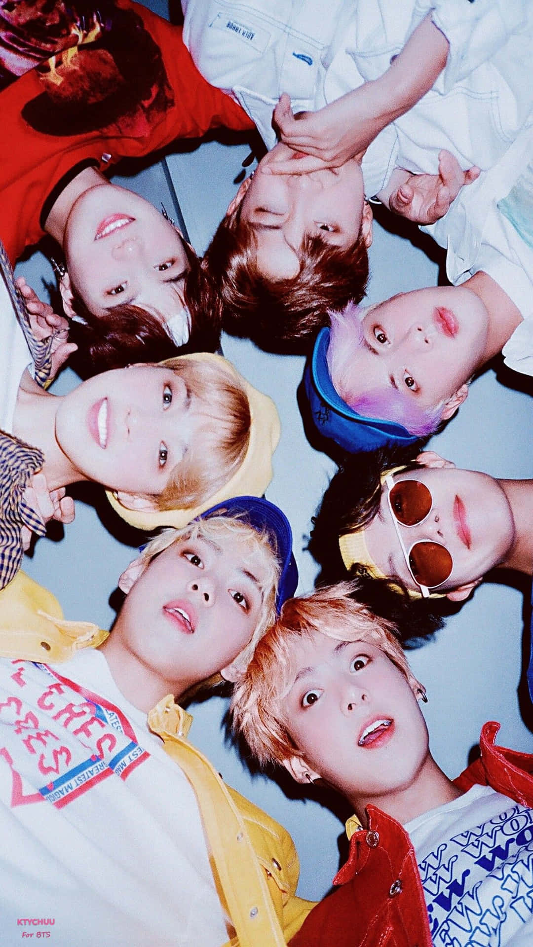 BTS臉部iPhone壁紙 壁紙 — 七名穿著色彩繽紛服裝的年輕男子在純藍色背景下緊密圍成一圈向上凝視。