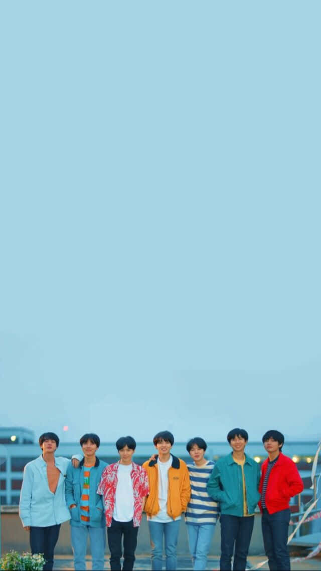 BTS藍天拍攝 壁紙 — 七個穿著彩色外套的年輕男子在黃昏時分站在屋頂上，背景是廣闊的淡藍色天空。