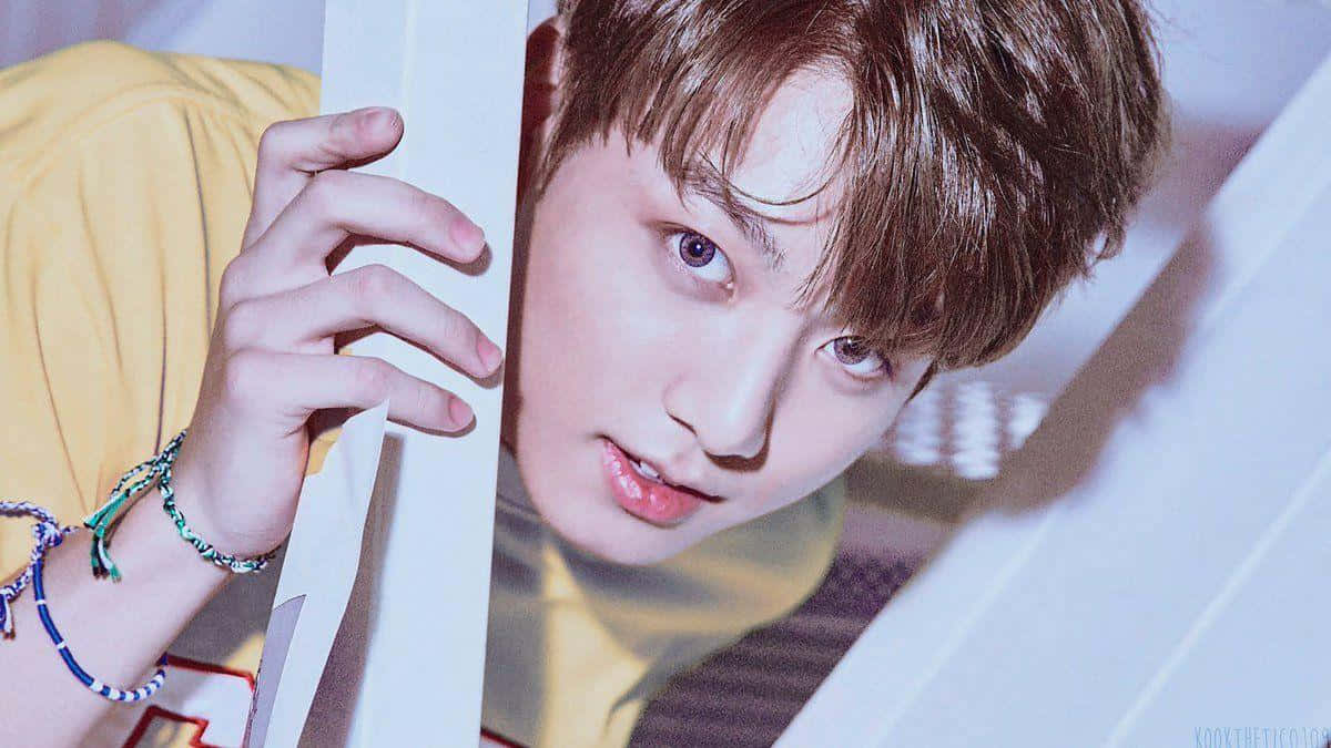 BTS超級巨星Jungkook舞台光芒四射 壁紙 — 一位棕色頭髮的年輕男子穿著黃色襯衫和彩色手鐲，透過白色板條屏障窺視。