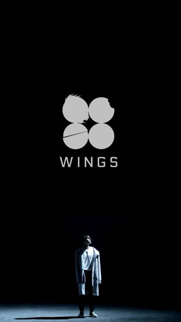 一個身穿白色外套的人，在黑暗的舞台上站在一個發光的「WINGS」標誌下方。