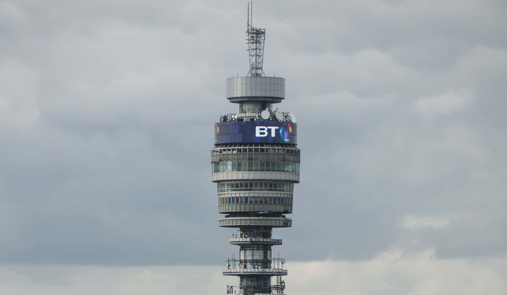 BT塔在多雲天空下 壁紙 — BT Tower 高聳入雲，其標誌性的圓柱結構和發光的藍色BT標誌在陰天背景下格外醒目。